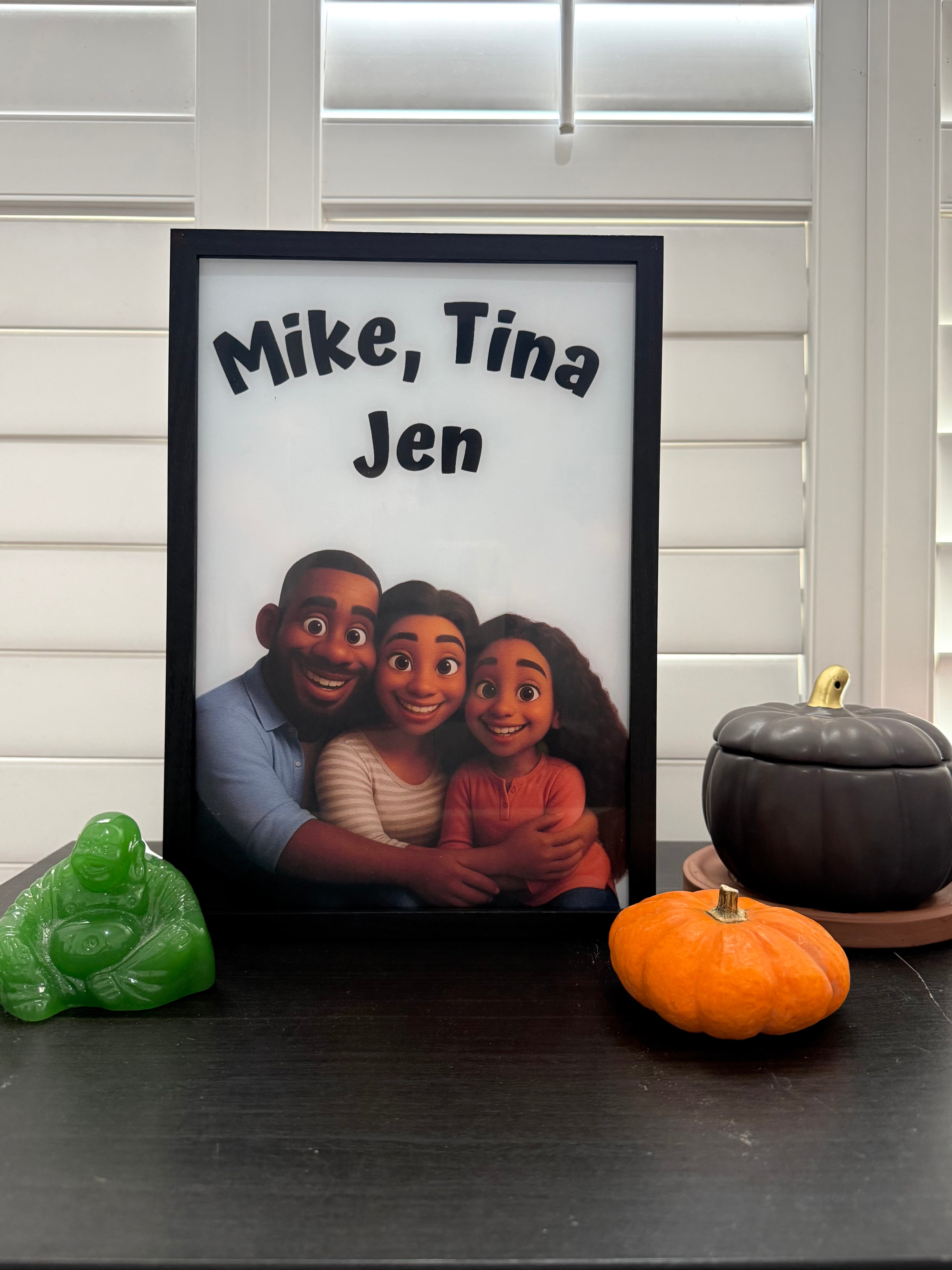 A framed custom photo print featuring a family portrait with the text 'Mike, Tina Jen' at the top, displayed on a dark tabletop with small decorative pumpkins and a green figurine in front of white shuttered windows.
